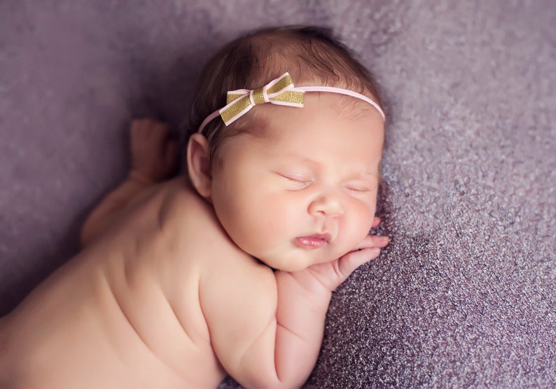 May include: A newborn baby girl sleeping on a purple blanket, wearing a pink and gold headband with a bow.