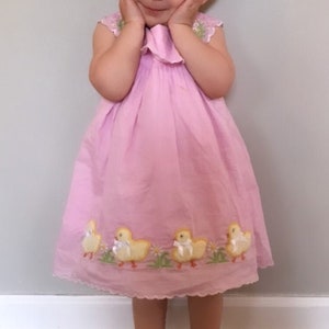 May include: A young girl wearing a pink dress with embroidered yellow chicks. She is smiling and has a blue bow in her hair.