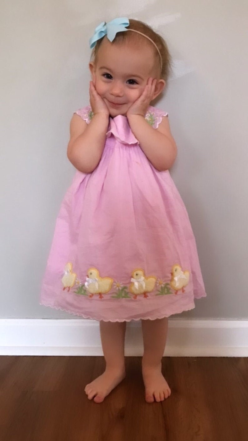 May include: A young girl wearing a pink dress with embroidered yellow chicks. She is smiling and has a blue bow in her hair.