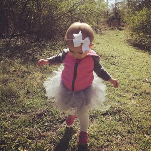 May include: A young girl wearing a pink jacket and a white tutu skirt with a white bow in her hair. She is walking on a grassy path.