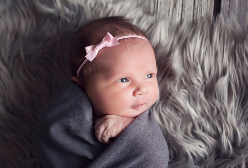 May include: A newborn baby wrapped in a gray swaddle, wearing a pink bow headband. The baby is lying on a gray fuzzy surface.
