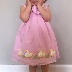 Pode incluir: Uma menina pequena vestindo um vestido rosa com pintinhos amarelos bordados. Ela est&aacute; sorrindo e tem um la&ccedil;o azul no cabelo.