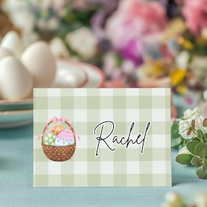 May include: A light green and white checkered place card with a watercolor illustration of a basket filled with decorated Easter eggs. The name "Rachel" is written in cursive.