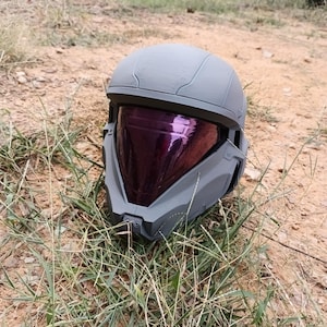 May include: A gray helmet with a purple visor rests on the ground. The helmet has a rounded top and angular features around the face. The visor is reflective, and the helmet appears to be made of plastic or a similar material.