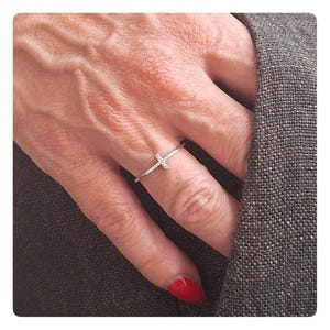 May include: A thin silver-toned ring featuring a small cross-shaped centrepiece adorned with tiny clear stones. The band is slender and metallic, designed to sit comfortably on a finger, showcasing a delicate and refined aesthetic for everyday wear.