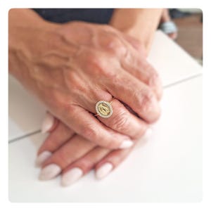 Peut inclure: Bague dorée ornée d'un pendentif ovale représentant une figure religieuse, entourée de petites pierres claires. La bague est montée sur un fin anneau. La bague est présentée sur les mains d'une personne aux ongles manucurés.