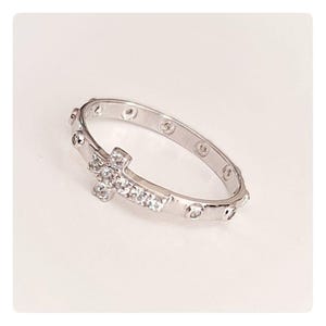 Puede incluir: Un anillo plateado con un diseño de cruz adornado con pequeñas piedras preciosas transparentes. La banda del anillo tiene pequeños detalles redondos. Este anillo es un accesorio religioso.