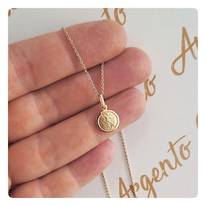 Op de afbeelding: Een goudkleurige ketting met een kleine, ronde hanger. De hanger heeft een gedetailleerd ontwerp en hangt aan een fijne ketting. De ketting wordt in een hand gehouden, wat de grootte en het ontwerp benadrukt.