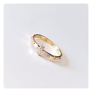 Puede incluir: Un anillo dorado con un diseño de cruz adornado con pequeñas piedras preciosas transparentes. El anillo tiene una banda con pequeños adornos redondos. El anillo está sobre un fondo blanco.
