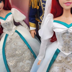 May include: A white wedding dress with a teal sash and silver embroidery. The dress has a sweetheart neckline and puffy sleeves. The doll is wearing a veil and has red hair.