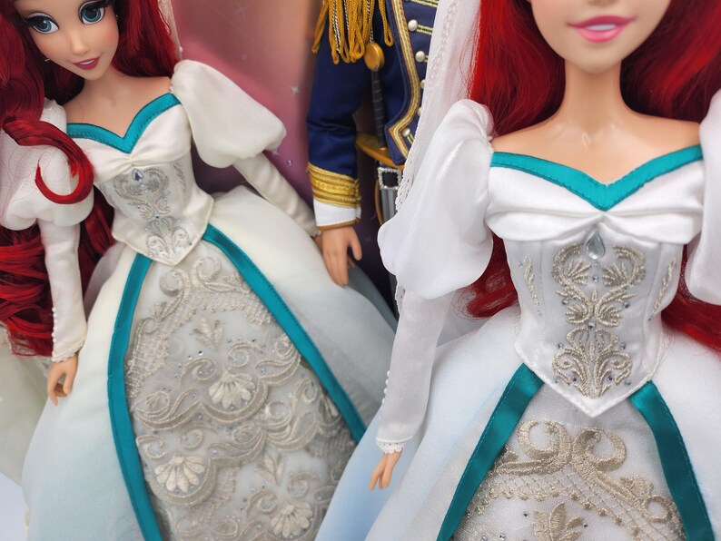 May include: A white wedding dress with a teal sash and silver embroidery. The dress has a sweetheart neckline and puffy sleeves. The doll is wearing a veil and has red hair.