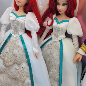 May include: Two Ariel dolls dressed in white wedding gowns with teal sashes and silver embroidery. The dolls have red hair and are wearing veils.