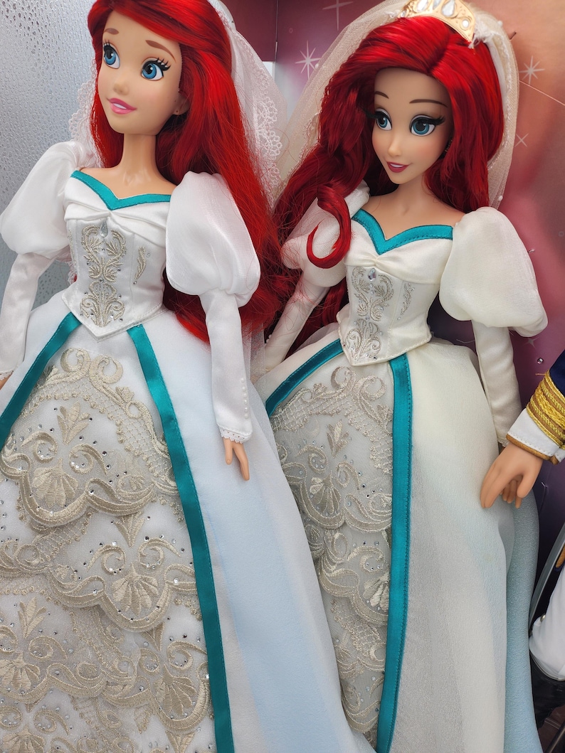May include: Two Ariel dolls dressed in white wedding gowns with teal sashes and silver embroidery. The dolls have red hair and are wearing veils.