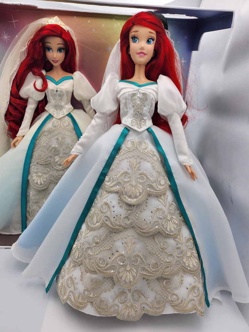 May include: A white wedding dress with a teal sash and gold embroidery. The dress has a long train and a fitted bodice. The doll is wearing a veil and has red hair.
