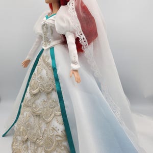 May include: A white wedding dress with a teal sash and gold embroidery. The dress has a long train and a veil with lace trim. The doll has red hair and is wearing a white veil.