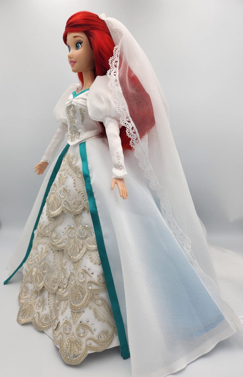 May include: A white wedding dress with a teal sash and gold embroidery. The dress has a long train and a veil with lace trim. The doll has red hair and is wearing a white veil.
