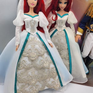May include: Two Ariel dolls dressed in white wedding gowns with teal sashes and gold embroidery. The dolls have red hair and are wearing veils.