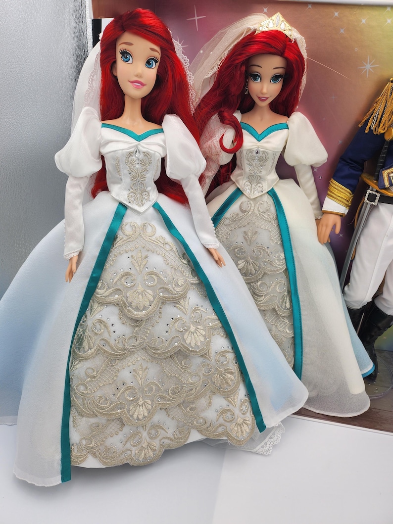May include: Two Ariel dolls dressed in white wedding gowns with teal sashes and gold embroidery. The dolls have red hair and are wearing veils.