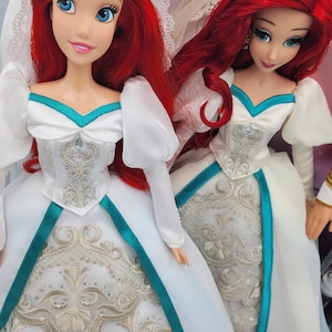 May include: Two Ariel dolls dressed in white wedding gowns with teal accents and silver embroidery. The dolls have long red hair and are wearing veils.