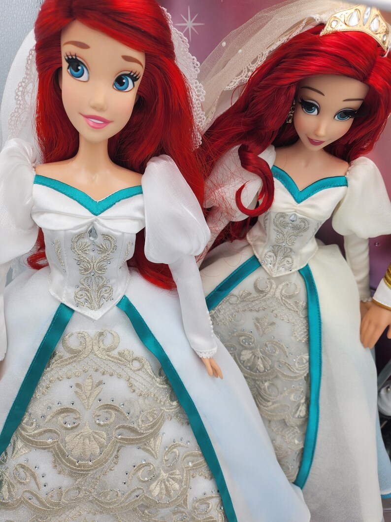 May include: Two Ariel dolls dressed in white wedding gowns with teal accents and silver embroidery. The dolls have long red hair and are wearing veils.