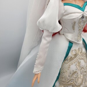 May include: A white wedding dress with a puffed sleeve, a teal sash, and gold embroidery. The dress has a long train and a veil.