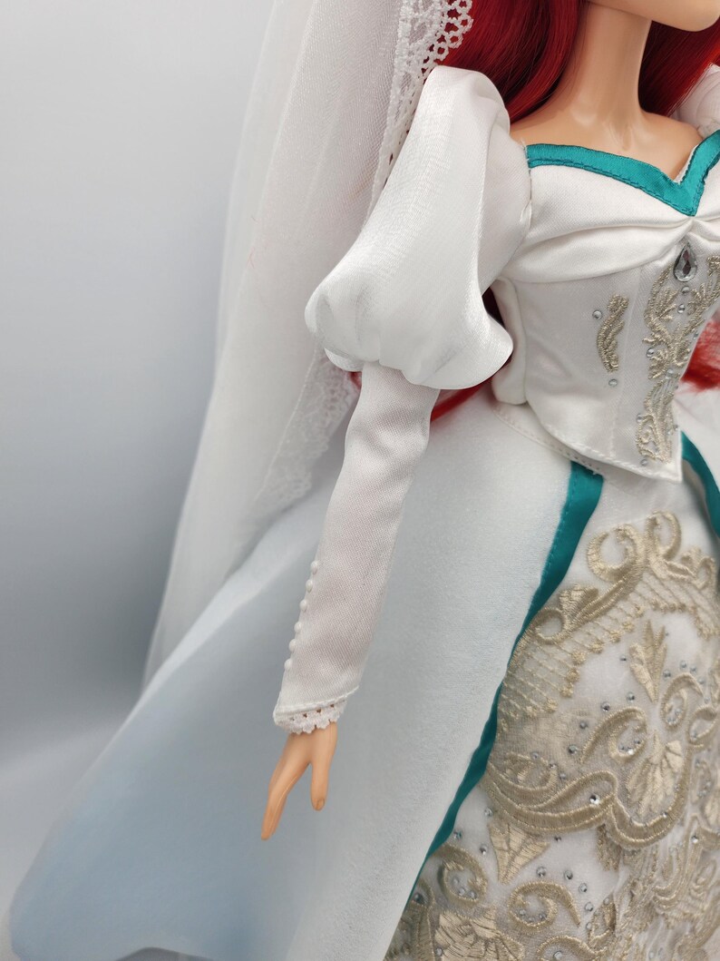 May include: A white wedding dress with a puffed sleeve, a teal sash, and gold embroidery. The dress has a long train and a veil.