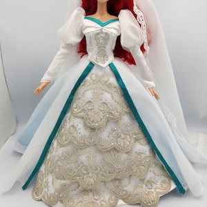 May include: A red-haired doll wearing a white wedding dress with gold embroidery and a teal sash. The dress has a long train and the doll is wearing a white veil.