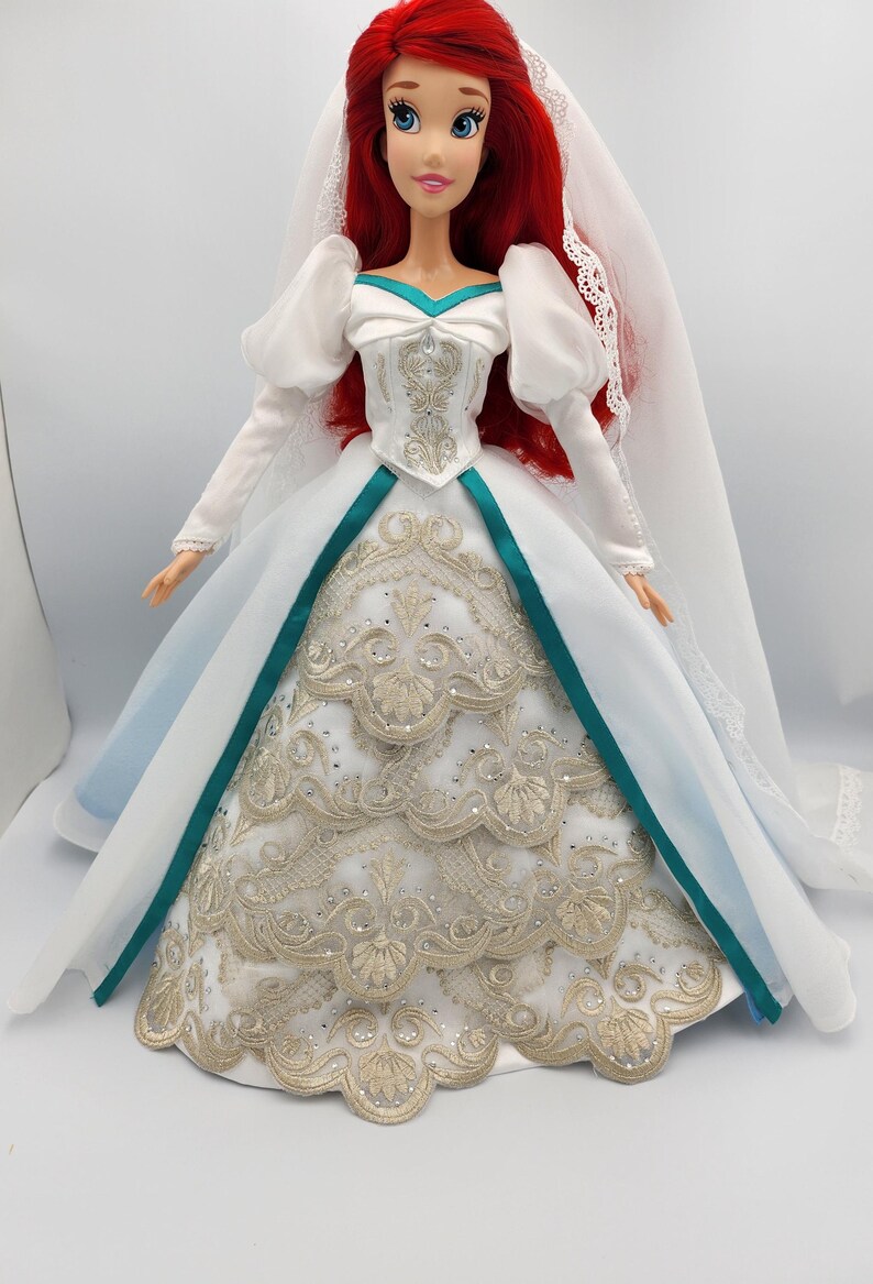 May include: A red-haired doll wearing a white wedding dress with gold embroidery and a teal sash. The dress has a long train and the doll is wearing a white veil.