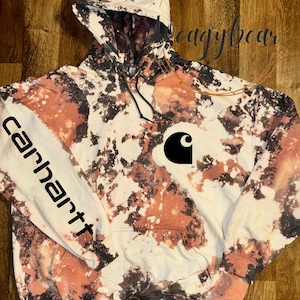 May include: A bleach-dyed hooded sweatshirt featuring a black and white mottled pattern. The garment displays a black Carhartt logo on the chest and the word carhartt printed vertically in black along the right sleeve. It includes a hood with drawstrings.