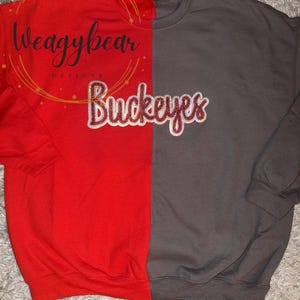 May include: A split-color crewneck sweatshirt featuring a bright red left side and a dark gray right side. The word "Buckeyes" is embroidered in a script font across the center in white with a red outline. A watermark reads "Weagybear DESIGNS".