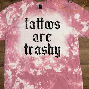 Könnte beinhalten: Ein kurzärmeliges T-Shirt mit Rundhalsausschnitt und einem rosa-weißen Batik-Bleichmuster. In der Mitte des T-Shirts befindet sich der schwarze Schriftzug "tattoos are trashy" in einer gotischen Schriftart auf einem gebleichten weißen Hintergrund.