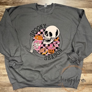 Può includere: Una felpa girocollo grigio antracite con una grafica circolare su sfondo a scacchi nero e rosa. Il design include un teschio che tiene in mano una tazza da caffè rosa, una piccola zucca con un cappello da strega e il testo SPOOKY SEASON.