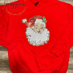 May include: A bright red long-sleeve sweatshirt featuring a large, central sequined graphic of a Santa Claus face. The Santa design includes a red hat with green holly accents and a white beard, all rendered in shimmering, textured sequins.