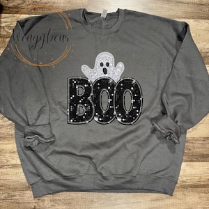 May include: A charcoal grey crewneck sweatshirt featuring a graphic design on the chest. The design includes a silver glitter ghost positioned above the word BOO, which is written in large, black, sequined block letters with a white outline.