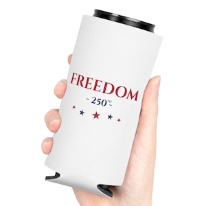 May include: White can cooler with the word "FREEDOM" in red and blue text. Below the word "FREEDOM" is the text "- 250"" and five stars. The can cooler is held in a hand.