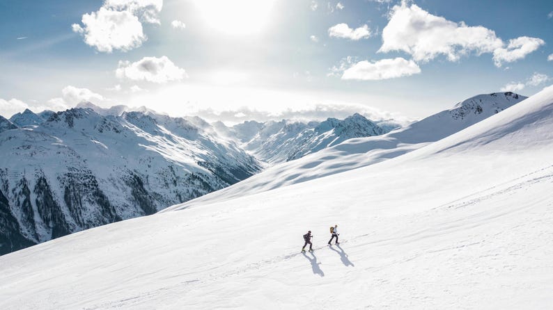 May include: Aerial view of two people hiking up a snow-covered mountain slope. The bright sun shines above, with a backdrop of snow-capped mountains and a partly cloudy sky. The scene captures a winter outdoor adventure.