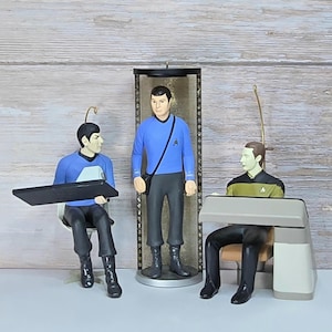 May include: Three Star Trek character figurines. One figure sits at a black console, another stands inside a cylindrical display case wearing a blue shirt with a black strap, and the third sits at a beige console wearing a yellow and black uniform.