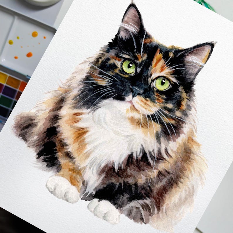 May include: Watercolor painting of a fluffy calico cat with green eyes. The cat has a mix of black, brown, and orange fur with a white chest and paws. The artwork is on white paper, with a watercolor palette visible in the background.