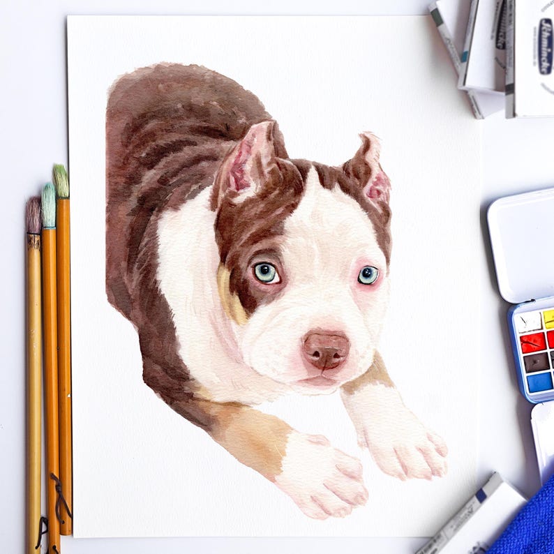 May include: Watercolor painting of a puppy with brown and white fur, and blue eyes. The puppy is lying down, looking towards the viewer. Paintbrushes and a watercolor palette are visible in the background.