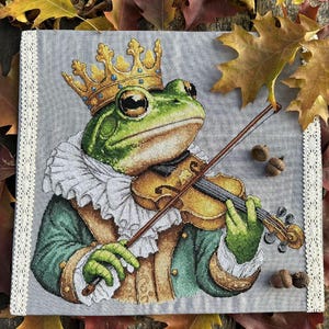 May include: Cross-stitch artwork of a regal frog playing a violin, wearing a gold crown and a ruffled collar. The frog is dressed in a green and gold coat. The artwork is framed with a white lace border, set against a backdrop of autumn leaves.