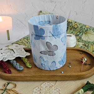 Puede incluir: Un recipiente de tela azul claro con un estampado de mariposas, exhibido en una bandeja de madera con suministros de bordado. La bandeja también contiene una vela encendida, tijeras y carretes de hilo.