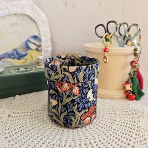 Puede incluir: Un recipiente cilíndrico de tela con un estampado floral en tonos azules, naranjas y crema. El recipiente está abierto en la parte superior y se apoya sobre una superficie blanca de ganchillo. Otros objetos están en el fondo.