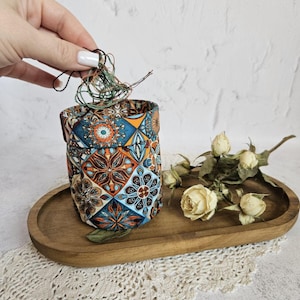 May include: A patterned fabric container with a drawstring closure, featuring a geometric design in shades of blue, orange, and brown. The container sits on a wooden tray with dried white roses.