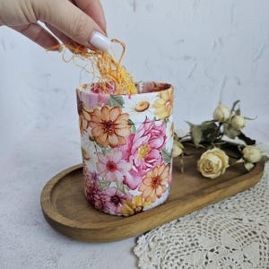 Puede incluir: Una cesta de almacenamiento cilíndrica de tela con un estampado floral vibrante de flores rosas, naranjas y amarillas sobre fondo blanco. La cesta contiene un pequeño ovillo de hilo de bordar naranja y descansa sobre una bandeja de madera ovalada.