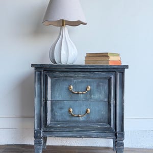 May include: A distressed blue bedside table with two drawers and brass handles. A white lamp with a scalloped shade sits on top, next to a stack of books. The table has a vintage, weathered appearance.