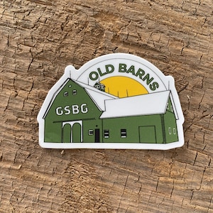 May include: A white sticker with a green barn illustration. The barn has a white roof and a yellow sun above it. The text "OLD BARNS" is in a circle above the barn. The text "GSBG" is on the side of the barn.