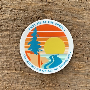 May include: A white round sticker with a sunset scene and a blue river. The text on the sticker reads "Meet me at the creek. I'm letting go of all my troubles."