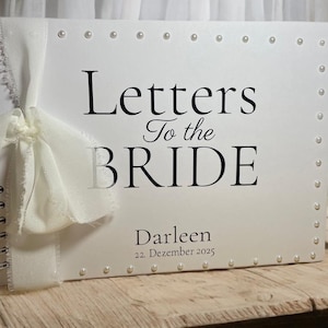 Könnte beinhalten: Ein weißes Gästebuch mit dem Text "Letters To the BRIDE" in schwarzer Schrift. Das Buch ist spiralgebunden und mit Perlen und einem weißen Band verziert. Der Name "Darleen" und das Datum "22. Dezember 2025" sind ebenfalls auf dem Cover aufgedruckt.