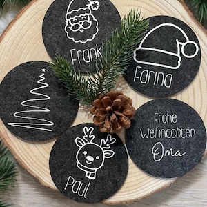Può includere: Cinque sottobicchieri natalizi in feltro grigio scuro con disegni bianchi. Ogni sottobicchiere presenta un design diverso: Babbo Natale, un albero di Natale, una renna, un cappello di Babbo Natale e il testo "Frohe Weihnachten Oma".