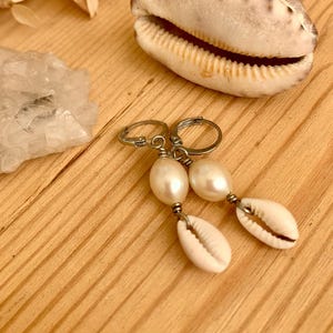 Könnte beinhalten: Ohrringe mit weißen Perlen und kleinen, weißen Muscheln, die an silberfarbenen Ringen befestigt sind. Eine große Muschel und Trockenblumen liegen auf einer Holzoberfläche. Ein Schmuckstück.